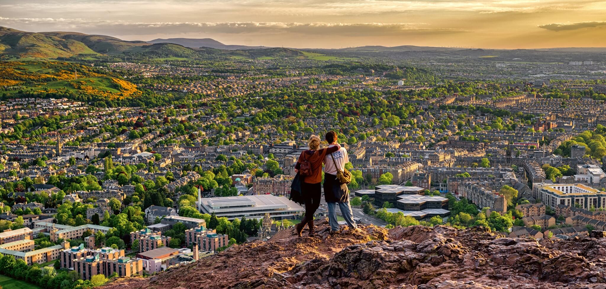 a couple standing at the top of arthurs seat in edinburgh at sunset overlooking the city